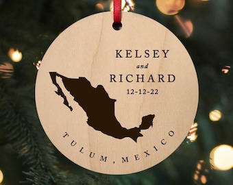 May include: A round, light-coloured wooden ornament hanging from a red ribbon. It features a dark brown silhouette of Mexico, the names KELSEY and RICHARD, the date 12-12-22, and the text TULUM, MEXICO printed in a dark, serif font on the front.
