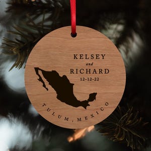 May include: A round wooden ornament featuring a dark silhouette of Mexico, the names KELSEY and RICHARD, the date 12-12-22, and the text TULUM, MEXICO. The ornament is suspended by a thin red ribbon through a hole at the top of the circular wood piece.