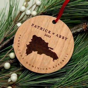 May include: A round wooden ornament featuring a laser-engraved map of the Dominican Republic, the text "PATRICK & ABBY 2023", and "PUNTA CANA, DOMINICAN REPUBLIC". The ornament is suspended by a thin red ribbon and has a natural light wood grain finish.