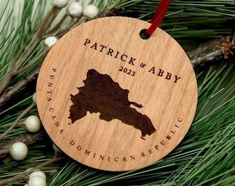 May include: A round wooden ornament featuring a laser-engraved map of the Dominican Republic, the text "PATRICK & ABBY 2023", and "PUNTA CANA, DOMINICAN REPUBLIC". The ornament is suspended by a thin red ribbon and has a natural light wood grain finish.
