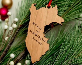 May include: A light brown wooden ornament shaped like the state of New Jersey, featuring the engraved text RYAN and MADISON with the date October 2023. The ornament is suspended by a thin red ribbon through a hole at the top of the wooden cutout.