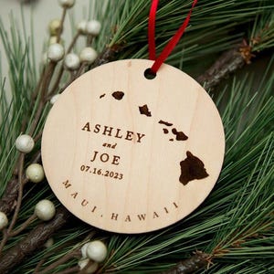 May include: A round, light-colored wooden ornament featuring a laser-engraved map of the Hawaiian Islands. The text reads ASHLEY and JOE, 07.16.2023, and MAUI, HAWAII. A thin red ribbon is threaded through a hole at the top for hanging purposes.