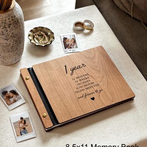 May include: A wooden memory book with the text "1 year" and related time measurements. The book is 8.5 x 11 inches and has a black spine. The image also shows several small photos, a decorative bowl, and some washi tape.