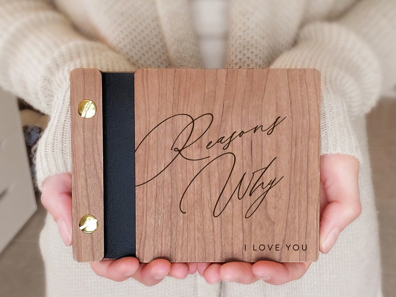 Two hands hold wooden engraved book with a close up of the cover. Reasons why I love you mini album with black binding and gold hardware.