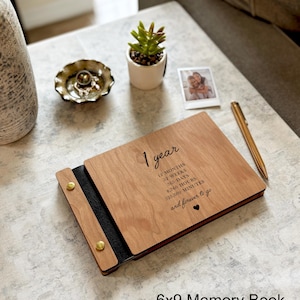 May include: A wooden memory book with the text "1 year" and time measurements. The book is open, revealing a black interior and gold-colored rivets. A pen and a small photo are next to the book. The text "6x9 Memory Book" is at the bottom.