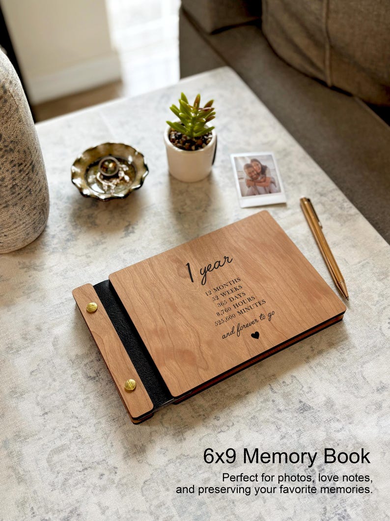 May include: A wooden memory book with the text "1 year" and time measurements. The book is open, revealing a black interior and gold-colored rivets. A pen and a small photo are next to the book. The text "6x9 Memory Book" is at the bottom.
