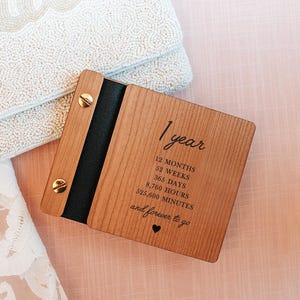 May include: A wooden keepsake book with the text "One Year. One Book." and "Designed for memories, notes, and love." The book is light brown with a black spine and gold-colored screws. The text on the book includes "1 year" and time measurements.