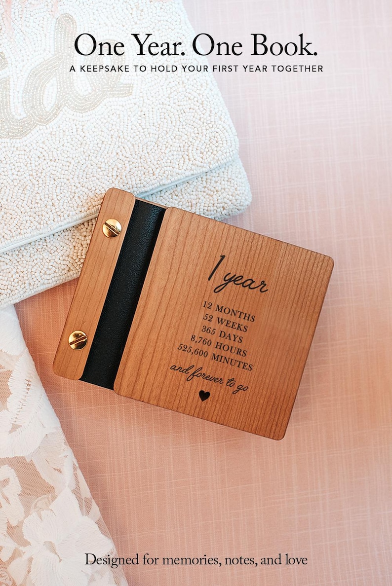May include: A wooden keepsake book with the text "One Year. One Book." and "Designed for memories, notes, and love." The book is light brown with a black spine and gold-colored screws. The text on the book includes "1 year" and time measurements.