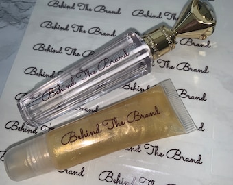 May include: Two clear lip gloss tubes with gold caps and the text "Behind The Brand" printed on them. The tubes are on a white background with multiple stickers that say "Behind The Brand".