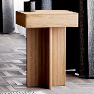 May include: A light wood end table with a simple, modern design. The table has a square top and four legs that are connected in the middle. The table is sitting on a light wood floor.