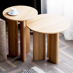 Puede incluir: Dos mesas auxiliares redondas de madera con un acabado en madera clara. Las mesas están anidadas y tienen un diseño simple y moderno. La mesa superior tiene una taza y un platillo blancos encima.