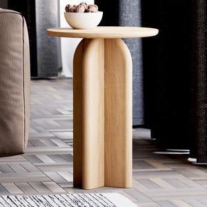 May include: A light wood side table with a round top and a unique, curved, open base. The table is in a living room setting with a brown sofa and curtains in the background.