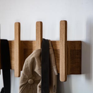 Könnte beinhalten: Eine hölzerne Wandgarderobe mit vier vertikalen Leisten, die an einer horizontalen Basis befestigt sind. Die hellbraune Holzmaserung ist auf der gesamten Struktur sichtbar. Ein schwarzer Riemen und eine beigefarbene Kappe hängen an den Haken.