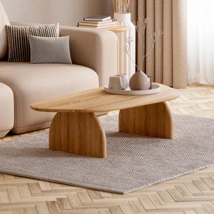 Op de afbeelding: Een lage, niervormige salontafel van lichtgekleurd hout met twee gebogen, massieve poten. Het tafelblad bevat een wit rond dienblad met een kleine keramische vaas en twee kleine kopjes, geplaatst voor een bank.