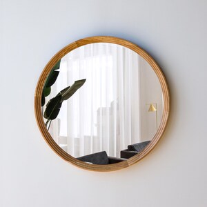 May include: A round mirror with a light brown wooden frame. The mirror reflects a room with a window, curtains, and a plant. The frame has a textured, ridged design. The mirror is mounted on a white wall.