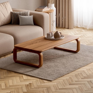 Könnte beinhalten: Ein niedriger, rechteckiger Couchtisch aus hellem Holz mit abgerundeten, durchgehenden Beinen. Auf der Tischplatte steht ein rundes Tablett mit zwei kleinen Keramiktassen und einer kleinen, abgerundeten Dekovase.