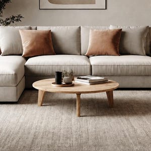 Puede incluir: Un salón moderno con un sofá seccional de color claro con cojines marrones y beige. Una mesa de centro redonda de madera se encuentra frente al sofá, y una gran pintura abstracta cuelga de la pared. La habitación tiene una paleta de colores neutros.