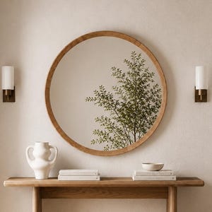 May include: A round wooden mirror hangs above a wooden console table. A white vase, books, and a small bowl sit on the table. Two sconces with white shades and brass bases flank the mirror. The mirror reflects a tree.