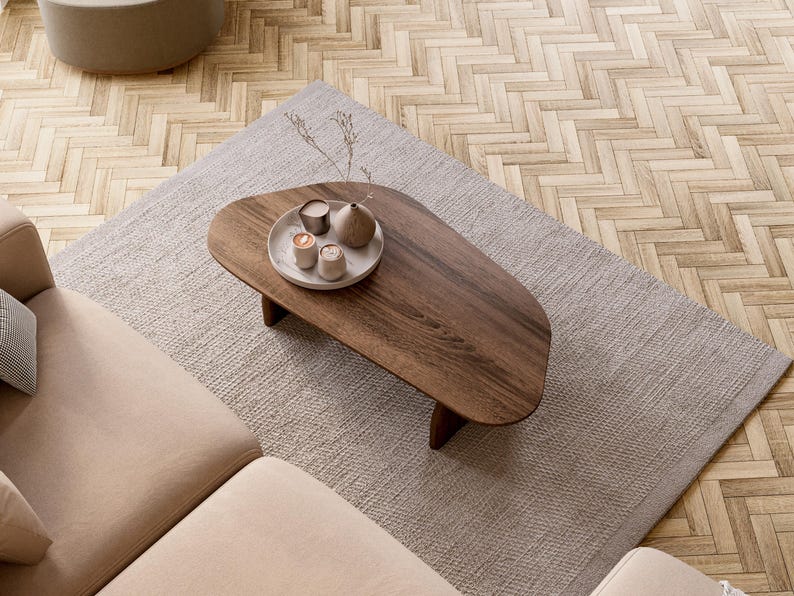 May include: An overhead shot of a living room setup featuring a dark wood coffee table with a decorative tray, a light gray rug, and a beige sofa. The table holds small candles and a vase. The floor is a light wood parquet.