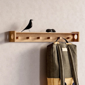 May include: A wooden wall shelf with six pegs, a black bird ornament, and sunglasses. A green and tan backpack hangs from the pegs. The shelf is made of light-coloured wood and is mounted on a neutral-coloured wall.