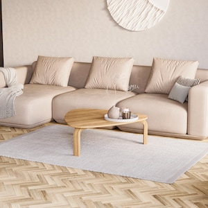 May include: A modern living room featuring a large beige sectional sofa with three large square pillows and a smaller patterned pillow. A light wood coffee table with a tray and decorative items sits on a light gray rug. A patterned throw blanket is draped over the sofa.