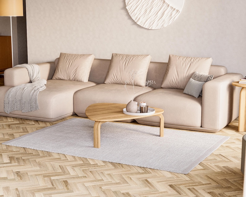 May include: A modern living room featuring a large beige sectional sofa with three large square pillows and a smaller patterned pillow. A light wood coffee table with a tray and decorative items sits on a light gray rug. A patterned throw blanket is draped over the sofa.