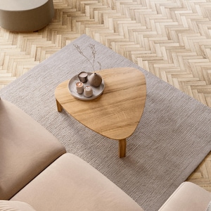 May include: An overhead shot of a living room setup featuring a light brown, heart-shaped wooden coffee table on a gray rug. The table holds a tray with small decorative items. A beige sectional sofa and a round ottoman are also visible.