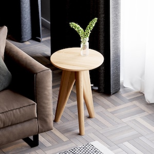 May include: A light brown wooden side table with a round top and three legs. The table is in a living room setting with a brown sofa and a vase of green flowers.