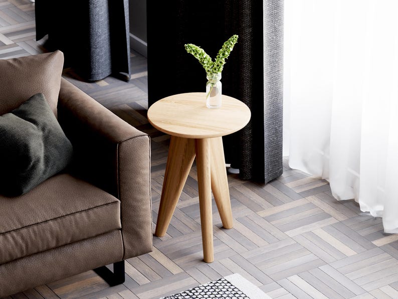 May include: A light brown wooden side table with a round top and three legs. The table is in a living room setting with a brown sofa and a vase of green flowers.