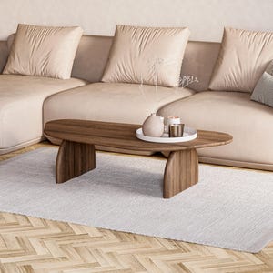 May include: A modern living room scene featuring a light beige sectional sofa with plush cushions. A wooden oval coffee table with decorative items sits on a white rug. The room has a neutral color palette.