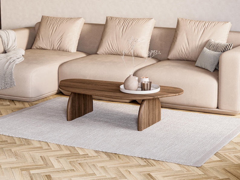 May include: A modern living room scene featuring a light beige sectional sofa with plush cushions. A wooden oval coffee table with decorative items sits on a white rug. The room has a neutral color palette.