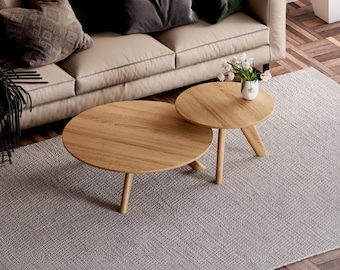 May include: A set of two round light oak coffee tables with smooth surfaces and tapered wooden legs. The tables are arranged in an overlapping configuration, with the smaller table holding a white vase containing white flowers.