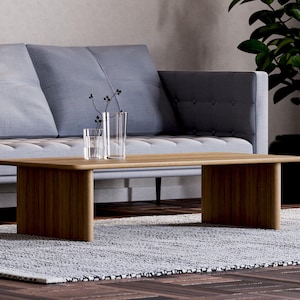 May include: A modern living room scene featuring a light wood coffee table with a rectangular top and two solid legs. A gray sofa with tufted cushions sits in the background. Two clear glass vases and a small glass are on the table. A potted plant is on the right.