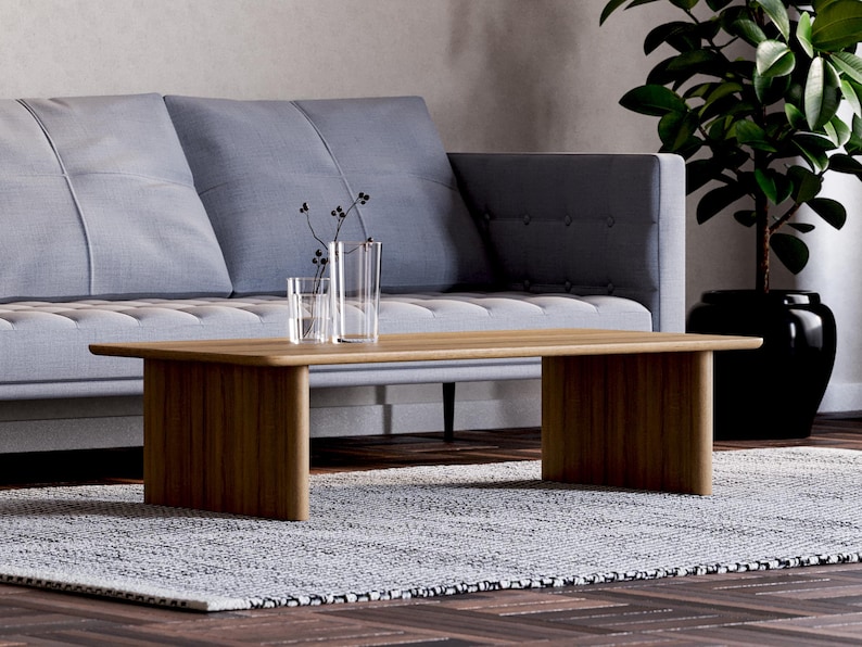May include: A modern living room scene featuring a light wood coffee table with a rectangular top and two solid legs. A gray sofa with tufted cushions sits in the background. Two clear glass vases and a small glass are on the table. A potted plant is on the right.