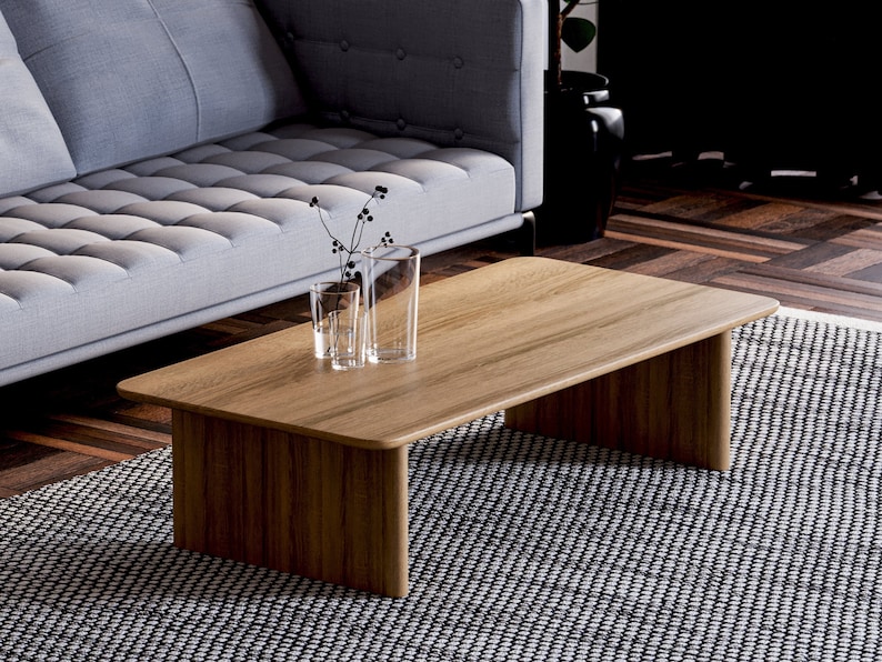 May include: A rectangular wooden coffee table with a light brown finish. The table has a simple, modern design with a flat top and two solid, rectangular legs. Two clear glass vases with decorative stems are on the table.