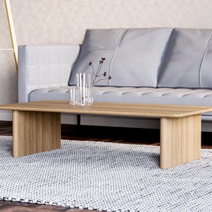 May include: A light brown rectangular coffee table with a light gray sofa in the background. The table has a simple design with a flat top and two rectangular legs. A clear vase and glasses are on the table. The rug is white with a black pattern.