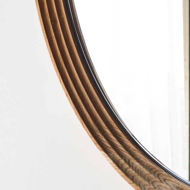 May include: Close-up of a wooden-framed mirror. The frame is made of light brown wood with a grooved texture. The mirror's edge has a dark, glossy trim. The background is a bright white.