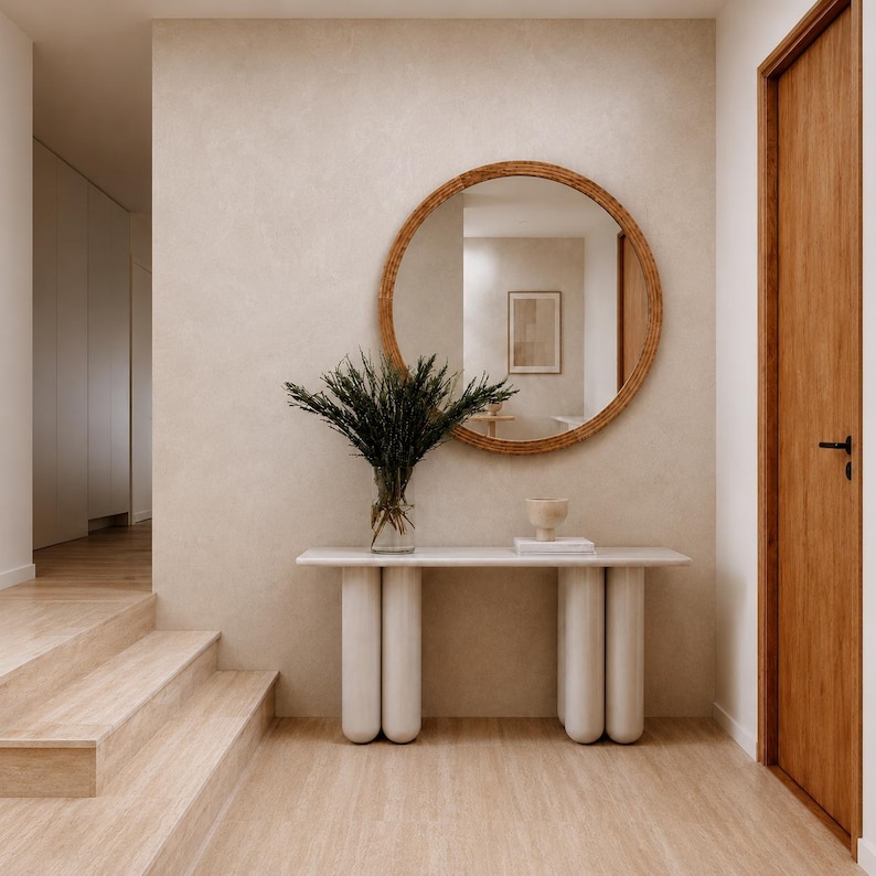 May include: A modern entryway featuring a round, woven-framed mirror above a white console table with cylindrical legs. A glass vase holds green foliage. The room has light-colored walls, wooden flooring, and a wooden door.
