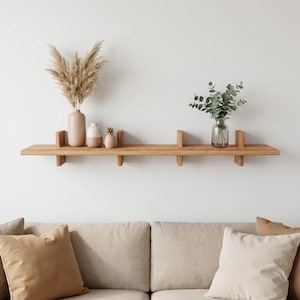 Peut inclure: Une longue étagère murale horizontale en bois avec quatre supports verticaux. À gauche, un grand vase beige contient de l'herbe de la pampa séchée, à côté d'un petit vase blanc et d'une figurine d'ananas. À droite, un vase en verre contient de l'eucalyptus.