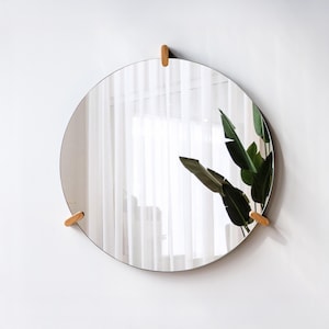 May include: A round mirror with a thin black frame, suspended by three small, light-colored wooden brackets. The mirror reflects a bright room with white curtains and a green plant. The design is minimalist and modern.