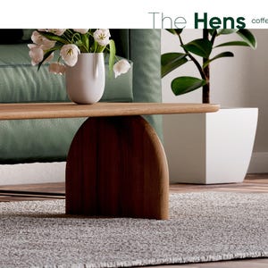 K&ouml;nnte beinhalten: Ein Holz-Couchtisch mit einem geschwungenen Sockel und einer hellen Holzoptik. Der Tisch steht auf einem grauen Teppich vor einem gr&uuml;nen Sofa.
