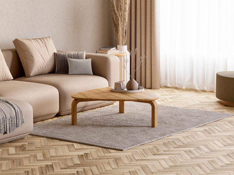 May include: A modern living room with a beige sectional sofa, a wooden triangular coffee table, and a gray rug. Decorative pillows and a vase with dried plants add to the room's aesthetic. The room has a neutral color palette.