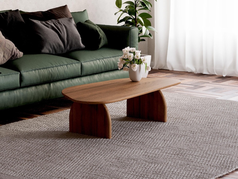 K&ouml;nnte beinhalten: Ein Holz-Couchtisch mit einer geschwungenen Oberseite und zwei Beinen auf jeder Seite. Der Tisch steht auf einem grauen Teppich vor einem gr&uuml;nen Sofa.