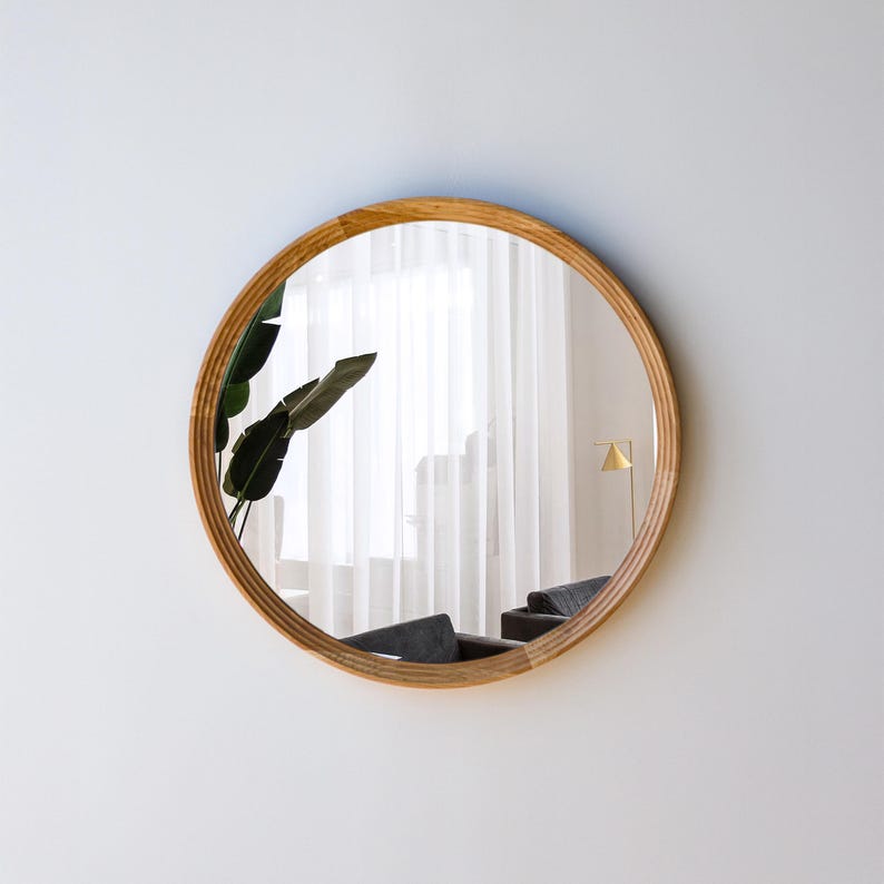 May include: A round mirror with a light brown wooden frame. The mirror reflects a room with a window, curtains, and a plant. The frame has a textured, ridged design. The mirror is mounted on a white wall.