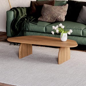 K&ouml;nnte beinhalten: Ein ovaler Couchtisch aus hellem Holz mit einem einzigartigen Design mit geschwungenen Beinen. Der Tisch steht auf einem wei&szlig;en Teppich vor einem gr&uuml;nen Sofa mit Kissen.