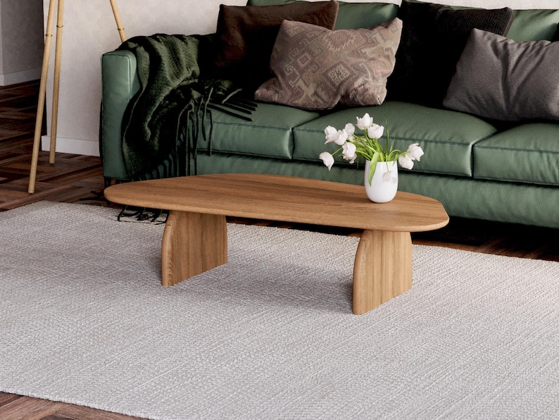 K&ouml;nnte beinhalten: Ein ovaler Couchtisch aus hellem Holz mit einem einzigartigen Design mit geschwungenen Beinen. Der Tisch steht auf einem wei&szlig;en Teppich vor einem gr&uuml;nen Sofa mit Kissen.