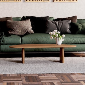K&ouml;nnte beinhalten: Ein gr&uuml;nes Sofa mit braunen und schwarzen Kissen, ein Holz-Couchtisch mit einer Vase mit wei&szlig;en Blumen und ein wei&szlig;er Teppich auf einem Holzboden.
