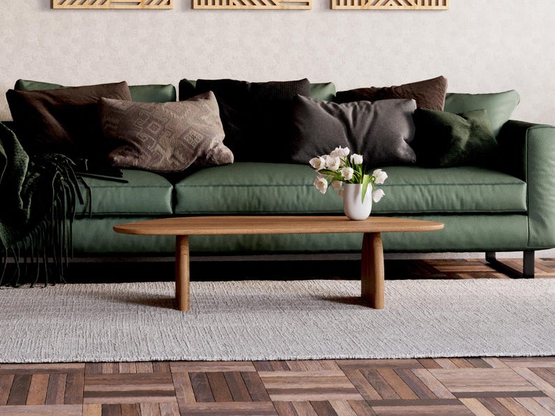 K&ouml;nnte beinhalten: Ein gr&uuml;nes Sofa mit braunen und schwarzen Kissen, ein Holz-Couchtisch mit einer Vase mit wei&szlig;en Blumen und ein wei&szlig;er Teppich auf einem Holzboden.