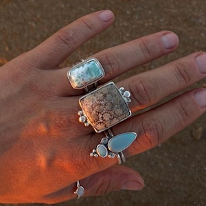 May include: Four silver rings worn on fingers. The index finger features a rectangular blue and white stone. The middle finger has a large square stone with a brown floral pattern. The ring finger displays three small blue stones. A thin band with a tiny stone sits on the pinky.