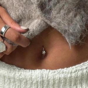 May include: A silver belly button ring with a small flower charm. The ring is worn on a person's navel. The person is wearing a grey sweater. The skin tone is light.